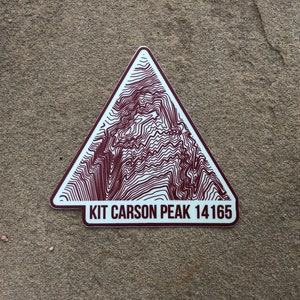 May include: A white triangular sticker with a brown topographic map design of a mountain peak. The sticker has the text "KIT CARSON PEAK 14165" printed on the bottom.