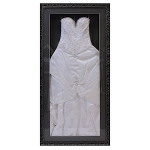 May include: A white strapless gown with intricate beadwork and embroidery on the bodice and skirt, displayed inside a tall, rectangular black frame with an ornate, textured border and a dark interior backing that highlights the dress details.