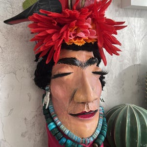 Puede incluir: Busto decorativo con rostro pintado, adornado con un pájaro rojo, flores y un collar de cuentas turquesas. El busto tiene largas pestañas y una corona de flores rojas. El busto está sobre una estantería de madera.