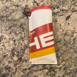 Puede incluir: Estuche de cuero blanco con un diseño a rayas rojo, amarillo y blanco. El estuche tiene una correa de cuero marrón y un cierre de presión metálico. El texto "HE" es visible en el estuche.