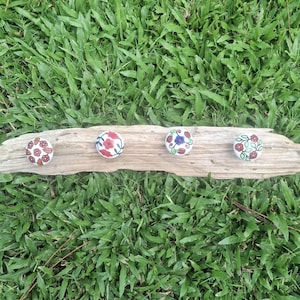 May include: A rustic wooden wall hook with four ceramic knobs. Each knob has a different floral design in red, brown, and blue on a white background. The wood is light brown, and the hook is set against green grass.