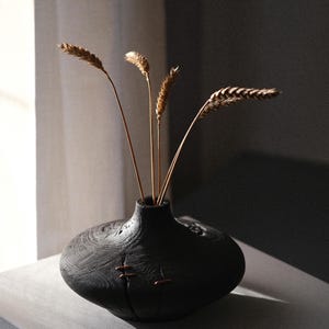 Pode incluir: Um vaso de madeira escura com uma forma arredondada única, segurando vários caules de trigo seco. O vaso tem uma superfície texturizada e detalhes em tons de cobre. O arranjo está sobre uma superfície cinza, iluminado por luz natural.