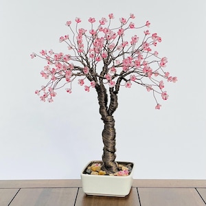 Op de afbeelding: Een handgemaakte bonsai-boomsculptuur met een gedraaide koperdraadstam en takken. De takken zijn versierd met delicate roze bloemenkralen. De boom staat in een witte vierkante keramische pot gevuld met kleine stenen.