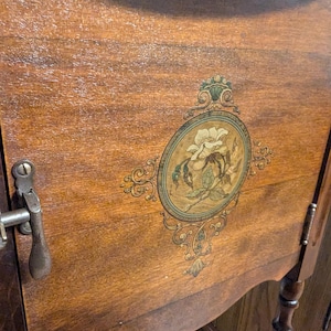 May include: A wooden cabinet with a single door and a floral design painted on the front. The door has a metal latch and the cabinet has turned legs.