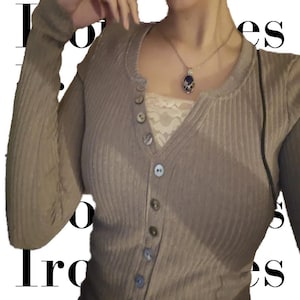 Puede incluir: Cárdigan de punto acanalado marrón claro con cuello redondo y cierre de botones. La parte superior de manga larga se combina con una camisola de encaje blanco y un collar plateado con un colgante azul. La palabra "Ironies" es parcialmente visible.