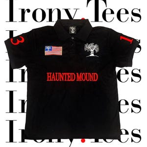Op de afbeelding: Een zwart poloshirt met rood geborduurde "HAUNTED MOUND"-tekst op de borst, een witte boomafbeelding en een kleine Amerikaanse vlag-patch. De mouwen tonen de rode cijfers 3 en 1, en de kraag is voorzien van een zwart label.
