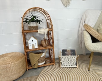 May include: A rattan shelf unit with three shelves, holding a potted plant, decorative items, and a woven basket. A macrame magazine rack and a round woven ottoman are also visible, creating a bohemian aesthetic.