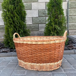 Puede incluir: Una cesta grande y ovalada de mimbre con dos asas. La cesta está tejida con juncos de color marrón claro y beige. La cesta está sobre una superficie de ladrillos grises, con dos árboles verdes al fondo.