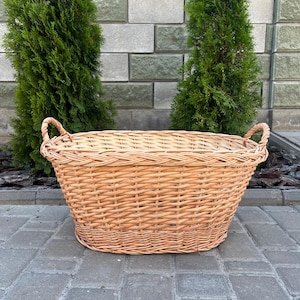 Puede incluir: Una cesta grande y ovalada de mimbre con dos asas curvas. La cesta está hecha de mimbre tejido de color marrón claro y se encuentra sobre una superficie de ladrillos grises. Dos árboles de hoja perenne verdes se encuentran en el fondo.