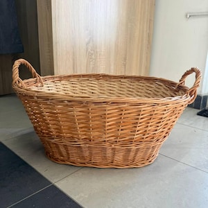 May include: A large, oval-shaped wicker basket with two sturdy handles. The basket is woven from light brown reeds, showcasing a natural, textured appearance. It sits on a tiled floor, suggesting its use for storage or display.