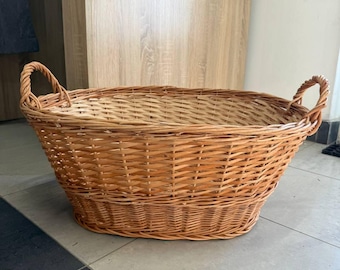 May include: An oval-shaped woven wicker basket featuring a natural light brown finish. The basket has a sturdy base, high sides with a cross-weave pattern, and two integrated handles on opposite ends for carrying and storage purposes.