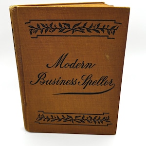 May include: A hardcover book titled Modern Business Speller with a tan fabric cover. The front cover features black text and decorative leaf borders at the top and bottom. The book is standing upright, showing the front cover and the spine edge.