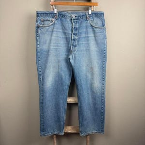 May include: Light blue denim jeans with a button fly and classic five-pocket design. The jeans are hanging on a wooden hanger. The denim has a faded appearance.