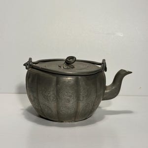 May include: Antique pewter teapot with a rounded body and a lid. The teapot features intricate floral and foliate designs etched into its surface. It has a curved spout, a handle, and a small loop on the lid. The teapot is a muted gray color.