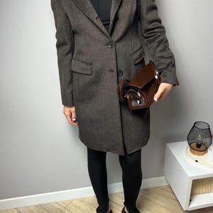 May include: A dark brown, knee-length coat with a two-button closure. The coat is paired with black leggings and brown shoes. A brown leather handbag is held in the hand. The outfit is set against a gray background.