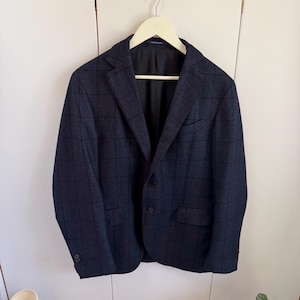 May include: A dark navy blue blazer featuring a subtle black windowpane check pattern. The jacket includes a notched lapel, two front buttons, a chest pocket, two flap pockets, and four buttons on each sleeve cuff, all hanging on a light hanger.