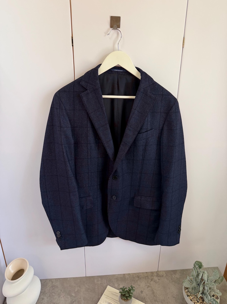 May include: A dark navy blue blazer featuring a subtle black windowpane check pattern. The jacket includes a notched lapel, two front buttons, a chest pocket, two flap pockets, and four buttons on each sleeve cuff, all hanging on a light hanger.