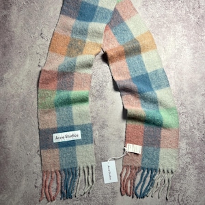 May include: A colorful scarf with a plaid pattern in pastel shades of pink, blue, green, and orange. The scarf has a fringed edge and a white label that reads "Acne Studios".