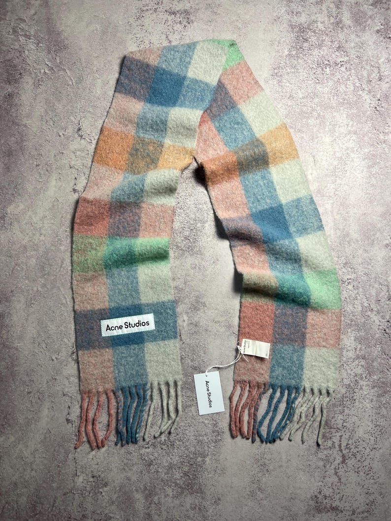 May include: A colorful scarf with a plaid pattern in pastel shades of pink, blue, green, and orange. The scarf has a fringed edge and a white label that reads "Acne Studios".