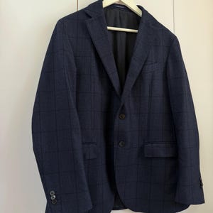 May include: A navy blue blazer featuring a subtle dark grid pattern, a notched lapel, two front buttons, and two flap pockets. The jacket has long sleeves with three small buttons on each cuff and is fully lined with a smooth black interior fabric.