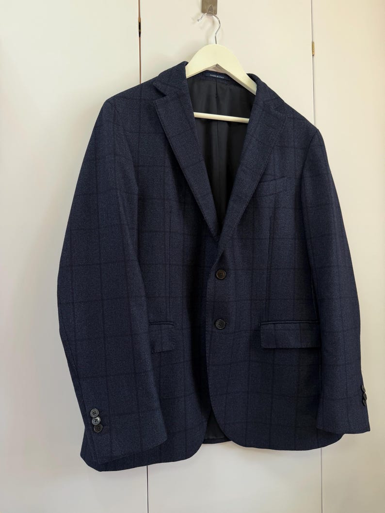 May include: A navy blue blazer featuring a subtle dark grid pattern, a notched lapel, two front buttons, and two flap pockets. The jacket has long sleeves with three small buttons on each cuff and is fully lined with a smooth black interior fabric.