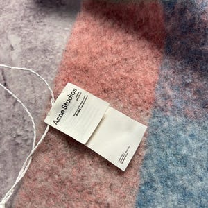 May include: A soft, colorful scarf with a plaid pattern in shades of pink, blue, and teal. The scarf has a textured, fuzzy appearance. A white tag with the text "Acne Studios" is attached.