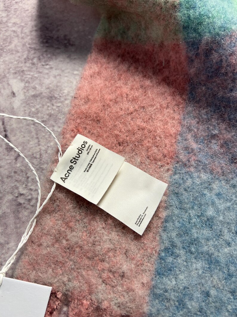 May include: A soft, colorful scarf with a plaid pattern in shades of pink, blue, and teal. The scarf has a textured, fuzzy appearance. A white tag with the text "Acne Studios" is attached.