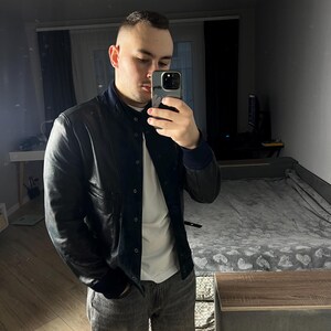 May include: A black leather jacket with a navy blue collar and cuffs. The jacket is worn over a white t-shirt and paired with gray jeans. The person is holding a phone, taking a mirror selfie.