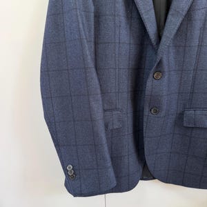 May include: A navy blue blazer featuring a dark grid pattern, a notched lapel, and two front buttons. The sleeves are finished with three small buttons at the cuffs, and the garment includes two flap pockets on the front and a black interior lining.