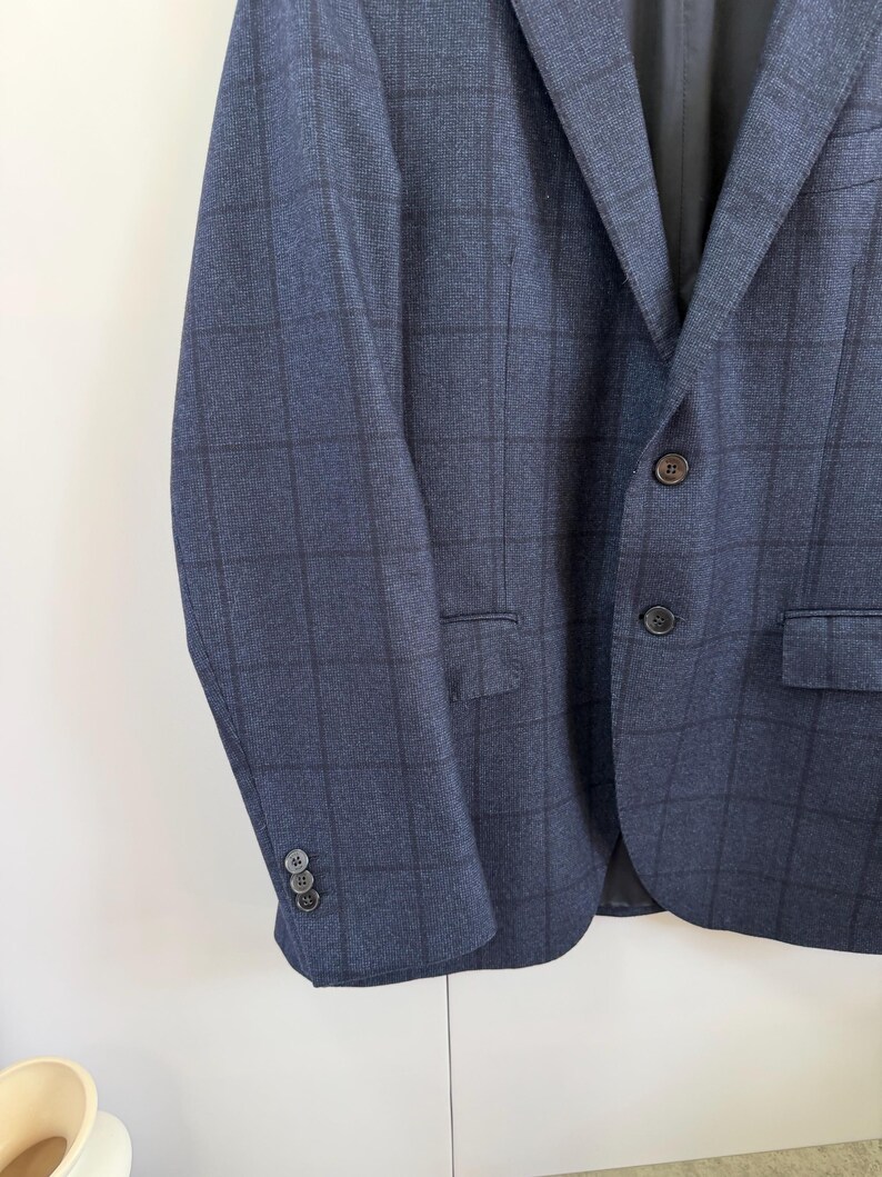 May include: A navy blue blazer featuring a dark grid pattern, a notched lapel, and two front buttons. The sleeves are finished with three small buttons at the cuffs, and the garment includes two flap pockets on the front and a black interior lining.