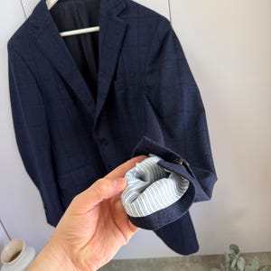May include: A dark blue blazer with a subtle grid pattern, featuring a notched lapel, a single-breasted front with buttons, and a sleeve cuff turned up to reveal a light blue and white striped lining on the interior of the garment.