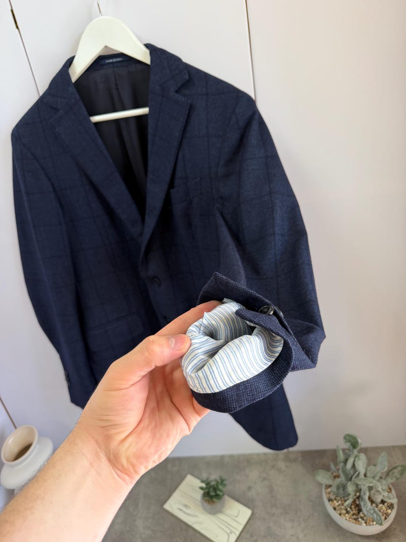 May include: A dark blue blazer with a subtle grid pattern, featuring a notched lapel, a single-breasted front with buttons, and a sleeve cuff turned up to reveal a light blue and white striped lining on the interior of the garment.