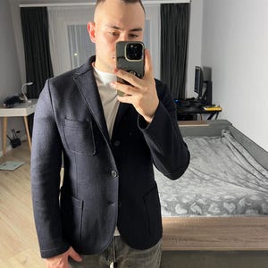 Peut inclure: Un blazer bleu marine foncé avec deux poches avant et deux boutons. Le blazer est porté sur un t-shirt blanc et associé à un pantalon gris. La personne tient un téléphone devant son visage.