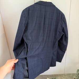 May include: A dark blue blazer featuring a subtle black grid pattern, a notched lapel, and a single vent at the back. The garment is constructed with long sleeves and a structured shoulder design, showcasing a classic tailored silhouette.