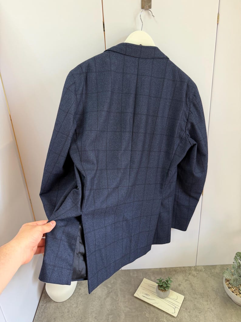 May include: A dark blue blazer featuring a subtle black grid pattern, a notched lapel, and a single vent at the back. The garment is constructed with long sleeves and a structured shoulder design, showcasing a classic tailored silhouette.