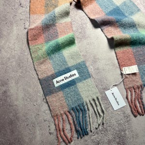 May include: A colorful scarf with a plaid pattern in shades of pink, blue, green, and orange. The scarf has a fringed edge and a white tag that reads "Acne Studios". The scarf is made of a soft, woven material.