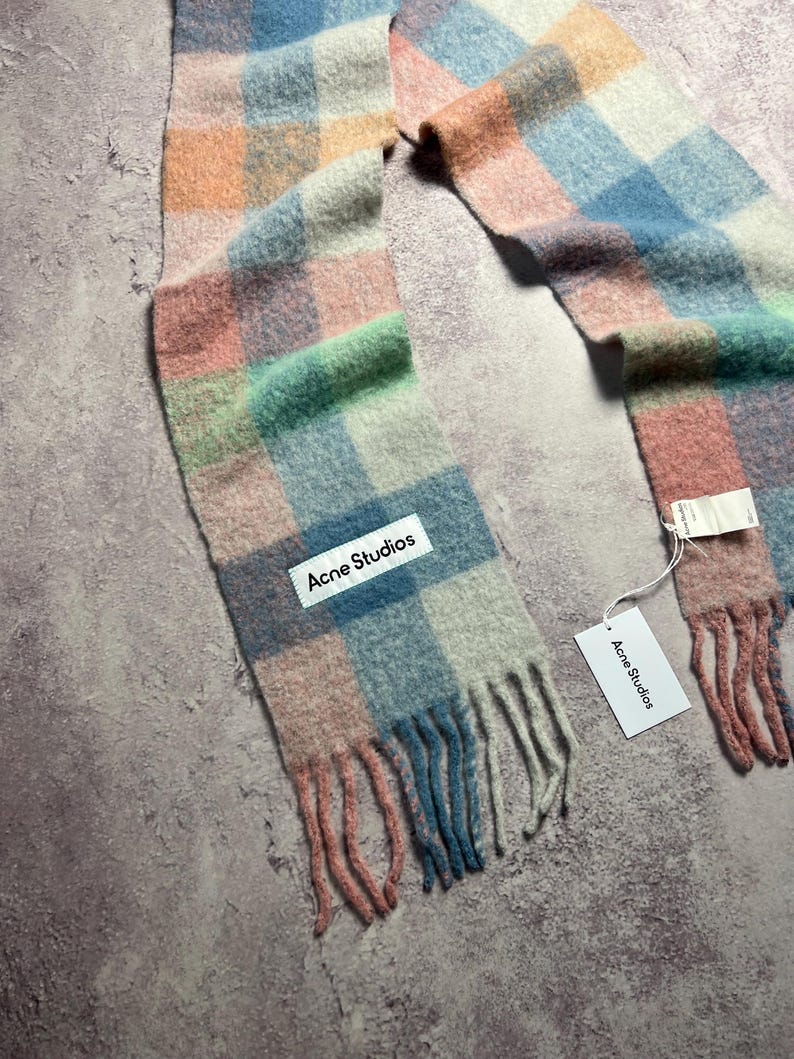 May include: A colorful scarf with a plaid pattern in shades of pink, blue, green, and orange. The scarf has a fringed edge and a white tag that reads "Acne Studios". The scarf is made of a soft, woven material.