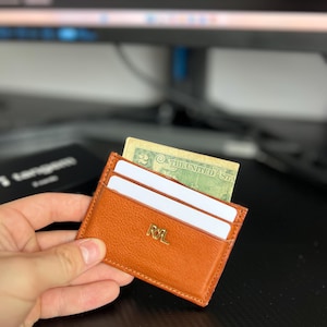 May include: An orange leather cardholder with the gold initials "RL" embossed on the front. The cardholder contains white cards and a US dollar bill. The cardholder is held in a hand.