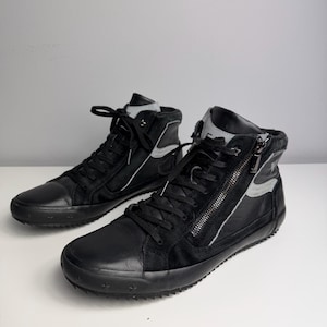 Könnte beinhalten: Paar schwarze High-Top-Sneaker mit Leder- und Wildleder-Einsätzen. Die Sneaker verfügen über einen seitlichen Reißverschluss, schwarze Schnürsenkel und einen grauen Akzent am Kragen. Die Sohlen sind schwarz und haben ein strukturiertes Design.
