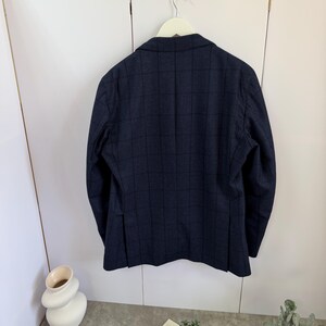 May include: A dark navy blue blazer featuring a subtle black windowpane check pattern. The jacket is displayed from the back, showing a structured collar, long sleeves, and two vertical vents at the bottom hem, hanging on a white hanger.