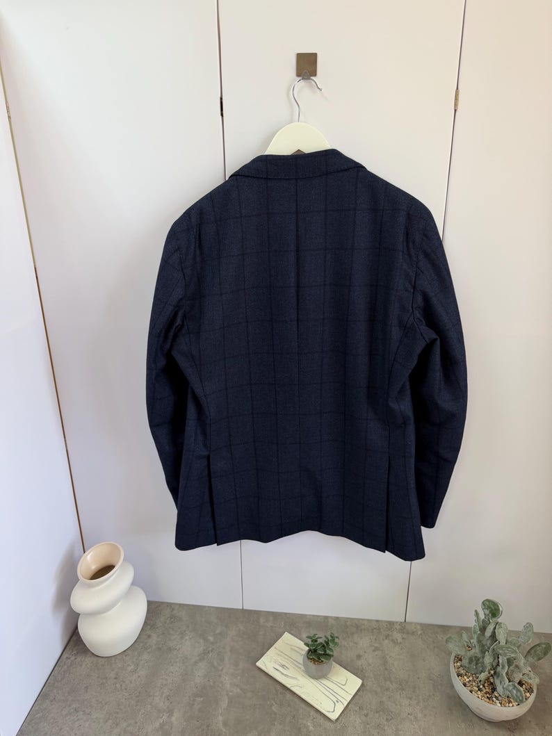 May include: A dark navy blue blazer featuring a subtle black windowpane check pattern. The jacket is displayed from the back, showing a structured collar, long sleeves, and two vertical vents at the bottom hem, hanging on a white hanger.