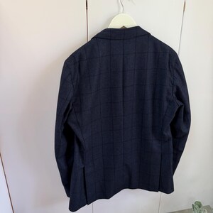 May include: A dark navy blue blazer featuring a subtle grid pattern in a darker shade. The blazer has long sleeves, a notched lapel collar, and a single vent at the back. It is displayed hanging on a white hanger against a plain white background.