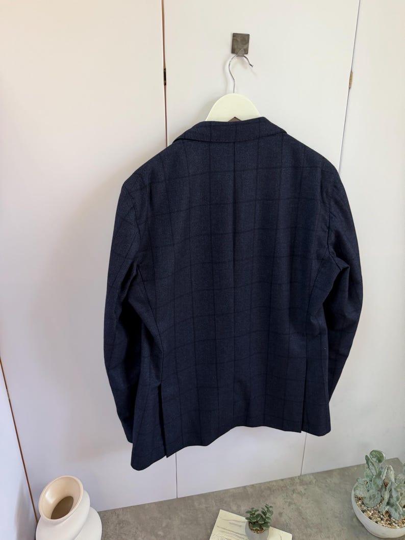 May include: A dark navy blue blazer featuring a subtle grid pattern in a darker shade. The blazer has long sleeves, a notched lapel collar, and a single vent at the back. It is displayed hanging on a white hanger against a plain white background.