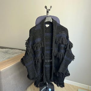 May include: A black and blue knitted cardigan with fringe detailing hanging on a hanger. The cardigan is on a garment steamer.