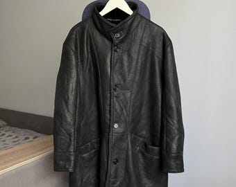 May include: A black leather coat with a button-up front and two front pockets hangs on a white hanger. The coat has a high collar and a classic design, suitable for cold weather.
