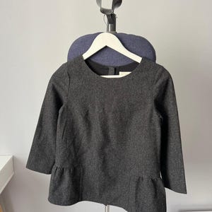 May include: A dark charcoal gray long-sleeved top featuring a round neckline and a peplum hem. The garment is constructed from a textured wool-like fabric and includes a black interior lining visible at the collar, with a small button closure at the back.