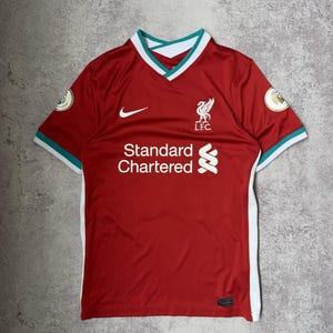 Op de afbeelding: Een rood voetbalshirt met een witte V-hals en turquoise accenten. Het shirt is voorzien van een witte Nike swoosh, het embleem van de Liverpool Football Club en het Standard Chartered-logo in het wit. Witte panelen lopen langs de zijkanten.