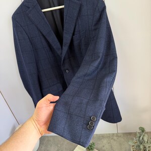 May include: A navy blue blazer featuring a dark grid pattern, a notched lapel, and a single-breasted front with a button closure. The sleeve cuff displays three small dark buttons. The interior lining is black, and a label reads Made in Italy.