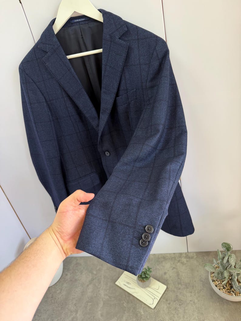 May include: A navy blue blazer featuring a dark grid pattern, a notched lapel, and a single-breasted front with a button closure. The sleeve cuff displays three small dark buttons. The interior lining is black, and a label reads Made in Italy.