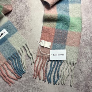 May include: A colorful, checkered scarf in pastel shades of pink, blue, green, and peach. The scarf has a fringed edge and a tag that reads "Acne Studios". The scarf is made of a soft, woven material.